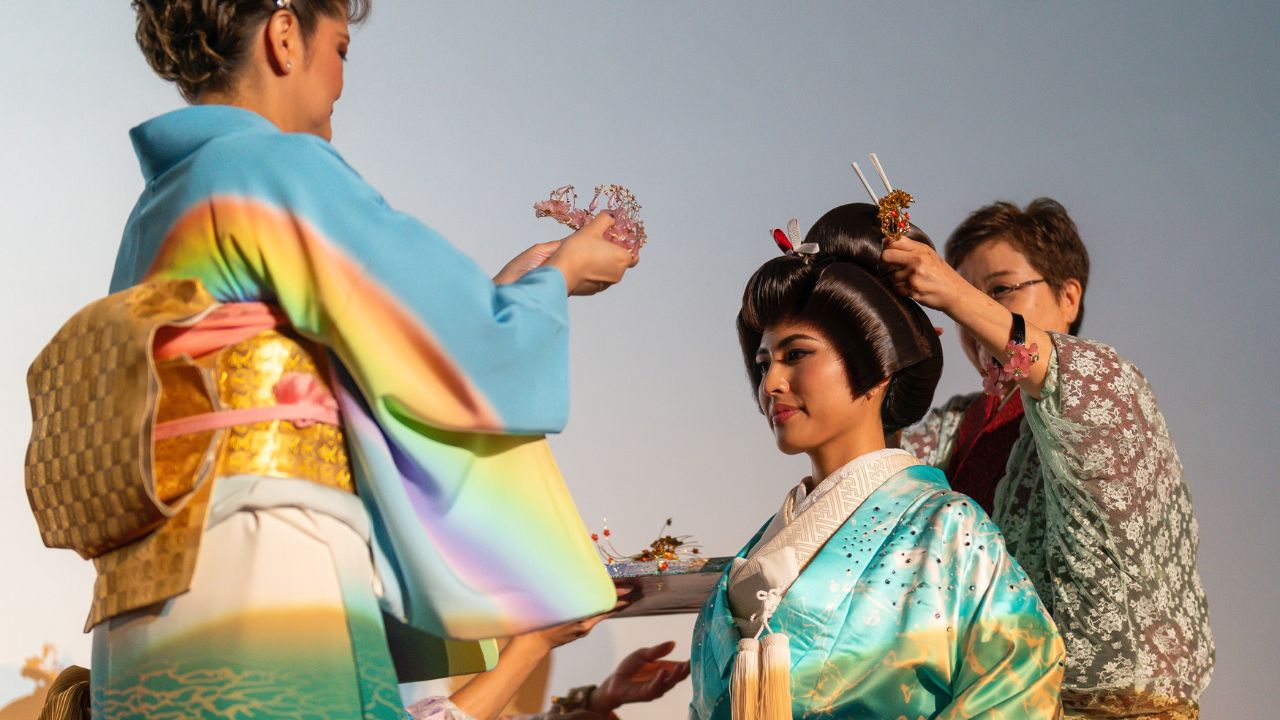 Jane Aiko Yamano and another woman in colorful traditional Japanese kimono help style another woman’s elaborate bridal hairstyle for wedding preparation.